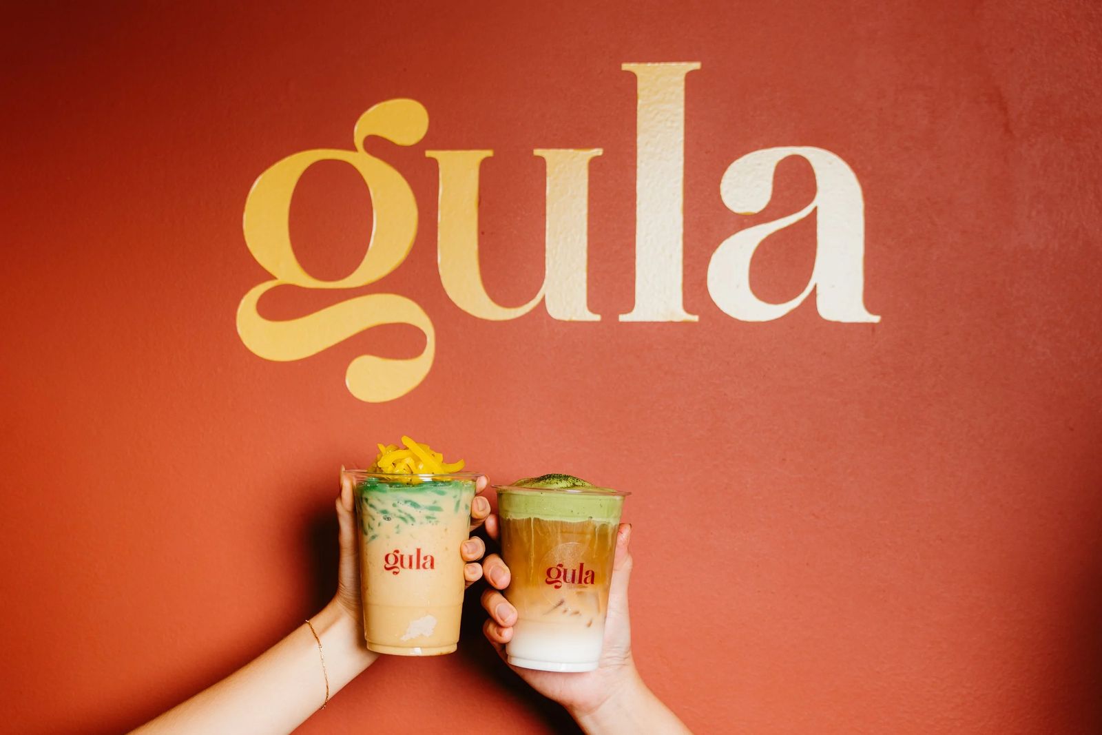 Two hands raising Gula drinks in front of the logo wall