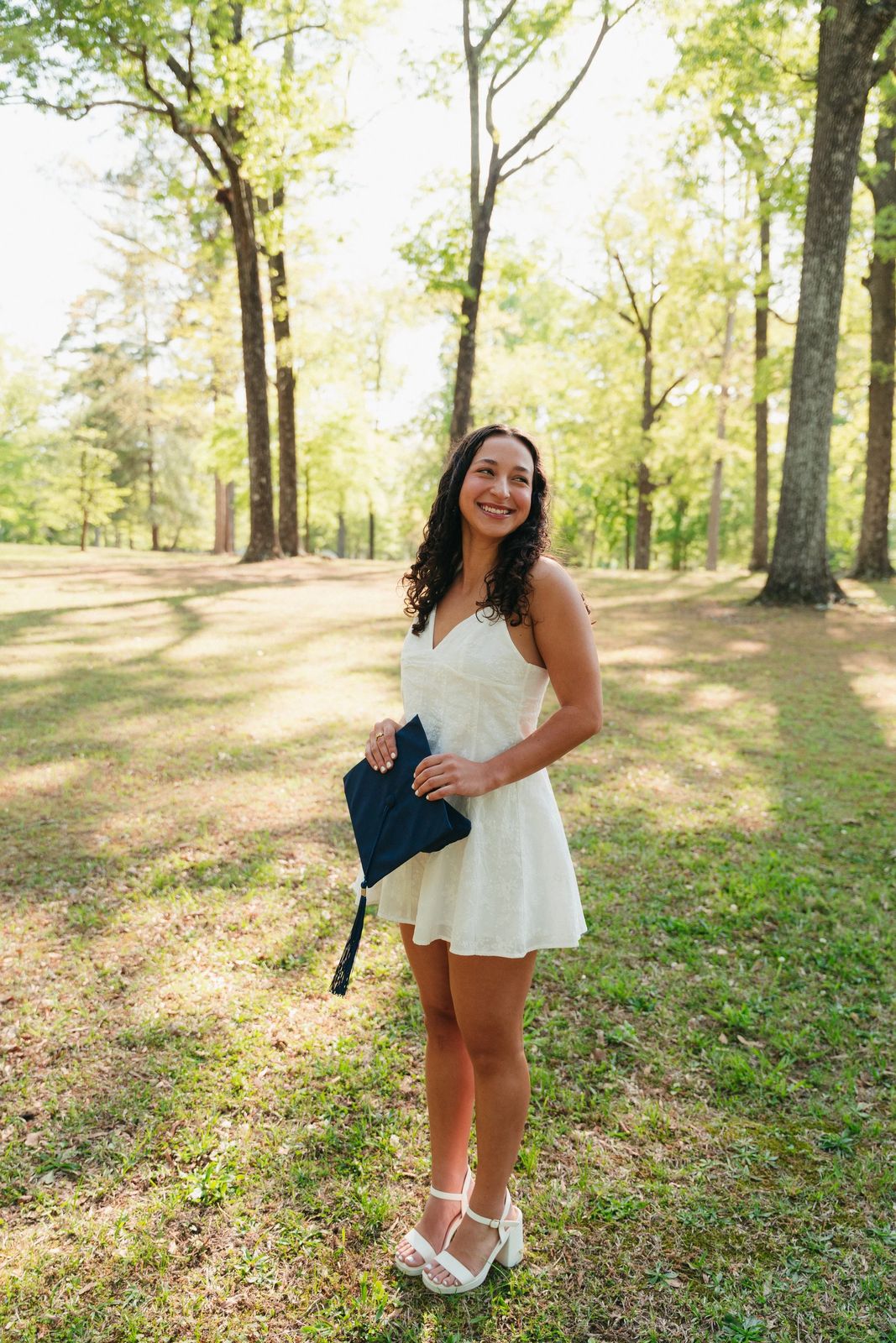 A senior holding her cap beneath tall trees